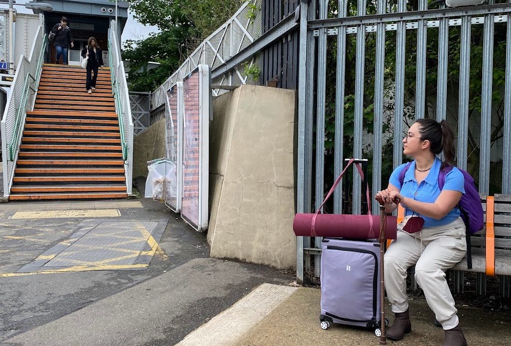 Traveler sitting on a bench with a cane, backpack, and rolling suitcase near stairs at a transit stop