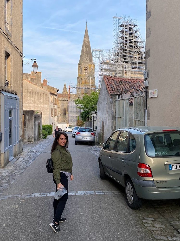 Monica Lynne, latina looking back on a quiet street in Le Dorat, rural France, with a church tower in the background
