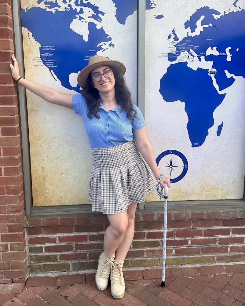 Monica Lynne, latina brunette leaning on brick wall in front of a window with white and blue picture of map behihnd her.
